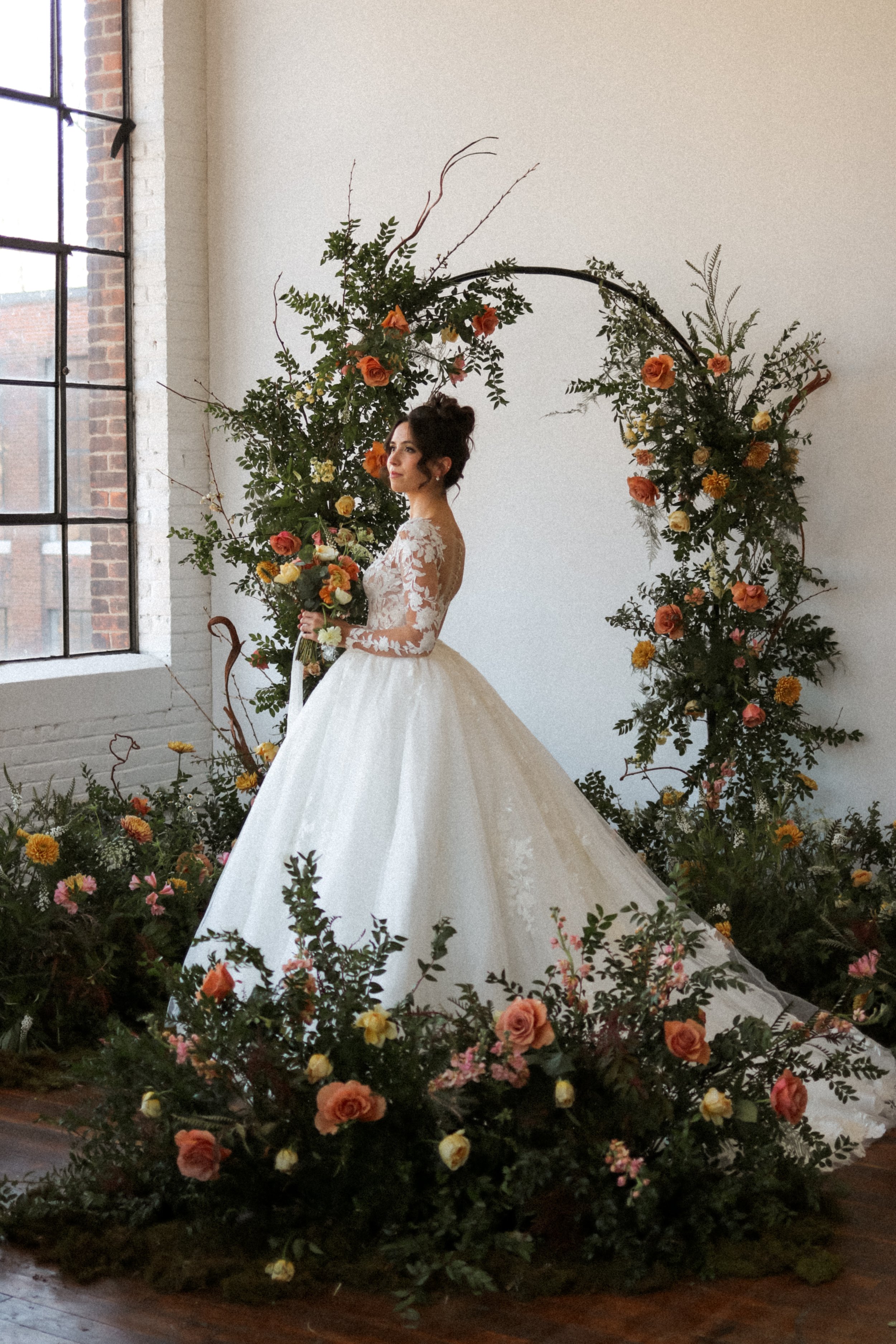 A bride in a white wedding dress holding a bouquet, standing in front of a floral wedding arch decorated with orange, yellow, and pink flowers, near a window in a bright room.